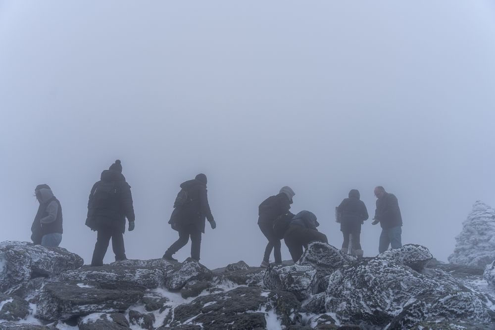 Silhouettes of hikers traverse a snowy mountain ridge in thick fog.