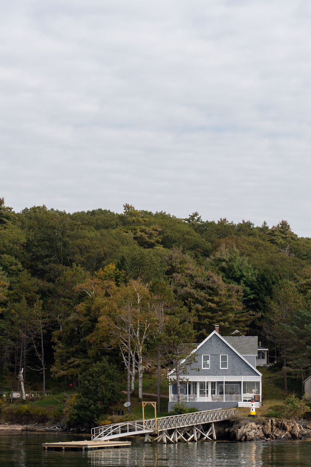 A grey waterfront cottage with a wraparound porch and wooden dock sits nestled among trees on a quiet shoreline.