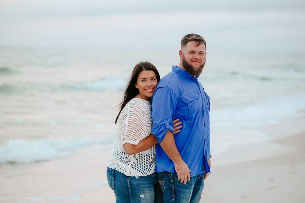 A couple embraces while standing on a sandy beach with ocean waves in the background.