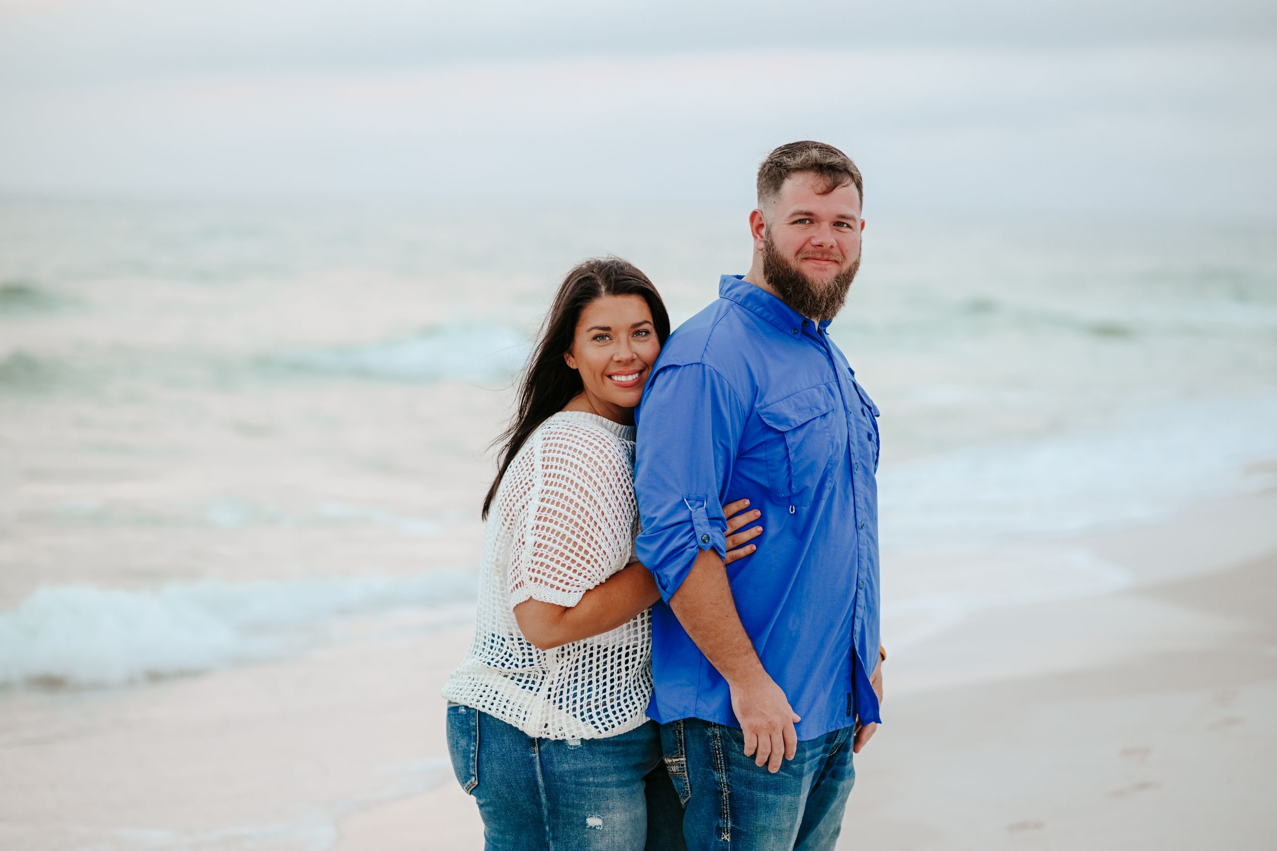 A couple embraces while standing on a sandy beach with ocean waves in the background.