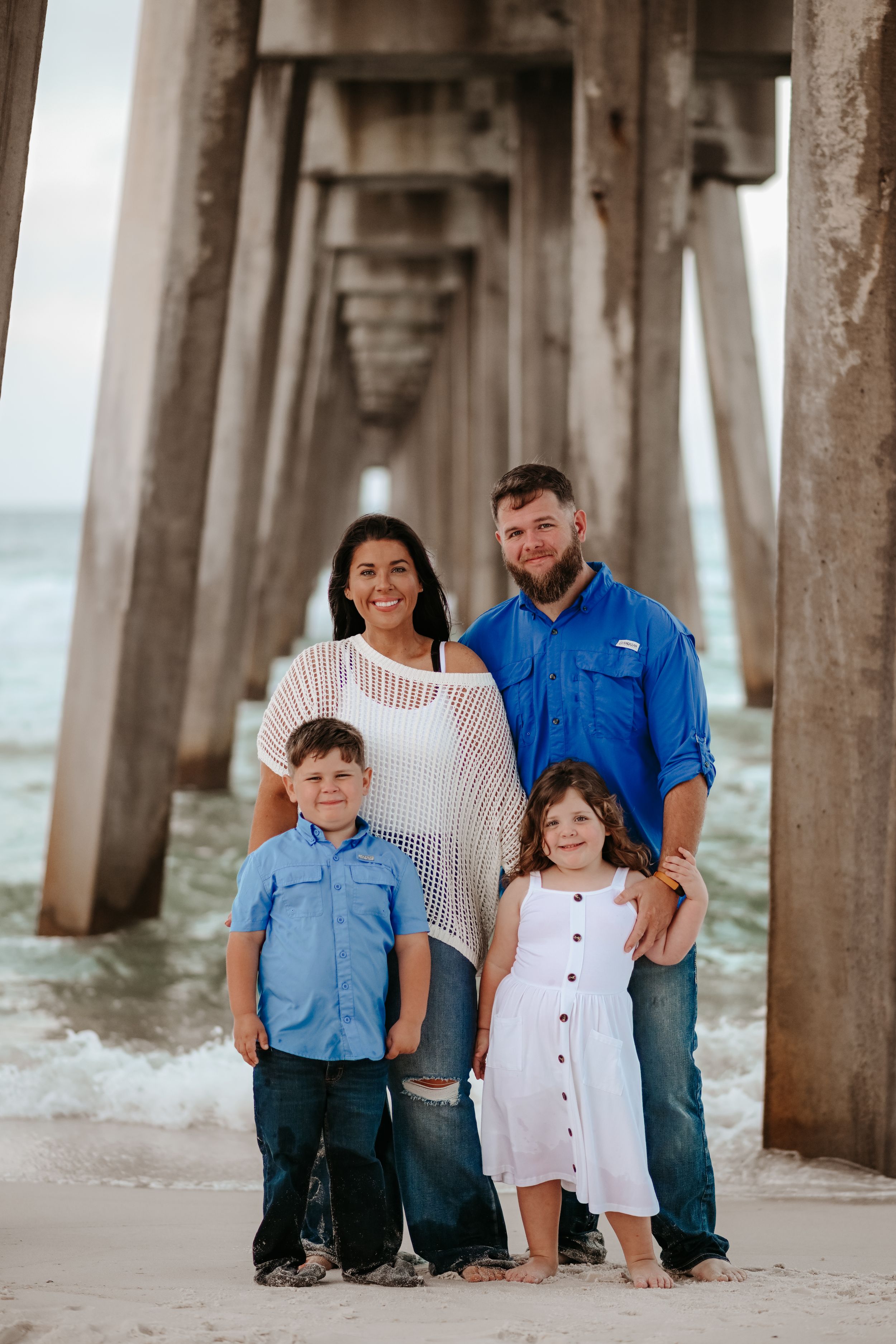 A family poses under a pier on the beach with waves crashing behind them.