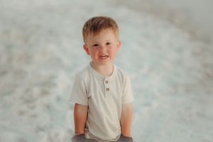 Child in white shirt sitting on sandy beach during photo session.
