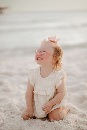 A young child wearing glasses and a white dress lies playfully in beach sand.