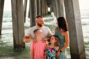 Family poses together under a wooden pier at the beach during golden hour.