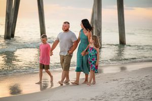 Family walks along shoreline under pier during sunset beach photoshoot.