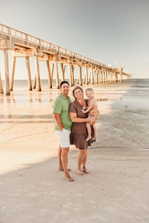 A family standing together on the beach with a long wooden pier in the background.