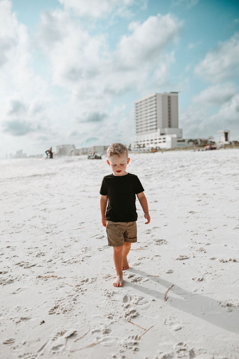 Small child walking alone on pristine white beach sand with coastal buildings visible in the background.