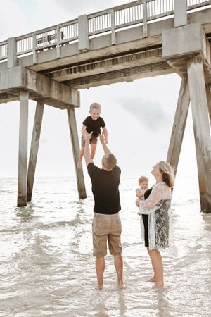 Family playing in the shallow water beneath a wooden pier on a cloudy beach day.