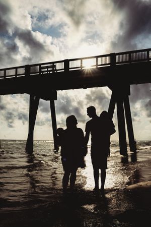 Dramatic silhouettes of people under a pier at sunset with moody clouds and ocean waves.