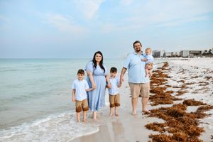 Family wearing coordinated blue outfits poses together on a sunny beach with seaweed and waves in the background.