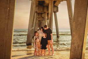 Silhouettes of people standing under pier supports at beach during golden hour.