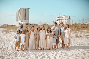 Large group wearing neutral colors poses for beach portrait with buildings in background.