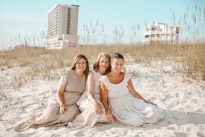 Three people sitting together on white sand dunes with coastal buildings in background.