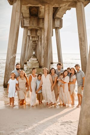 Large group in coordinated white and beige outfits posing together under a wooden pier at sunset on the beach.