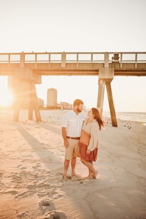 Couple standing beneath a wooden pier during golden hour at the beach.