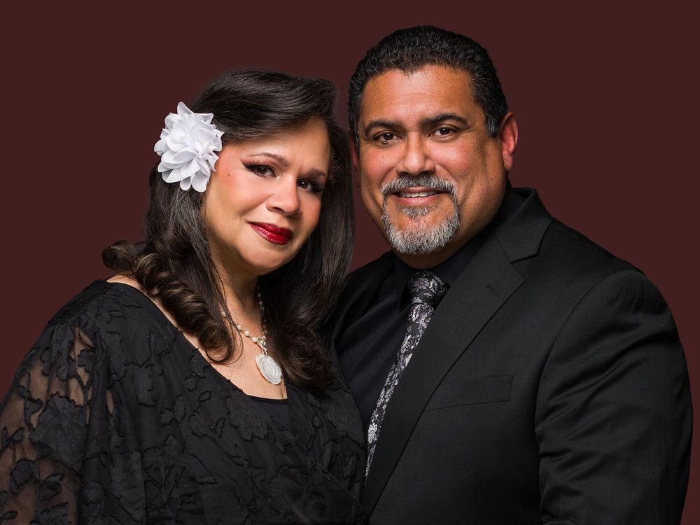 A couple in formal attire pose together, with the woman wearing black and the man in a dark suit.