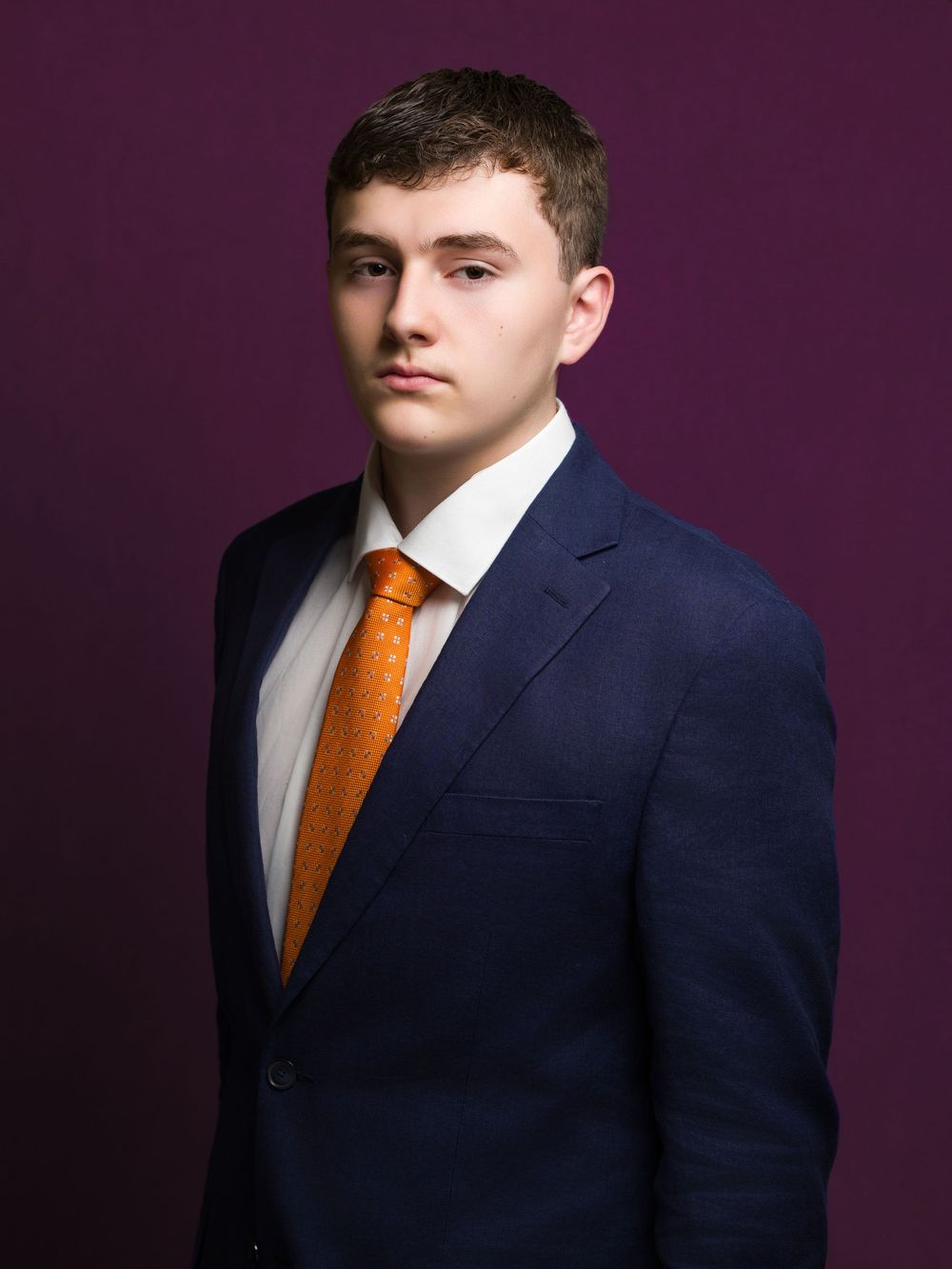 Professional headshot portrait of someone wearing a navy suit and orange tie against a dark purple backdrop.