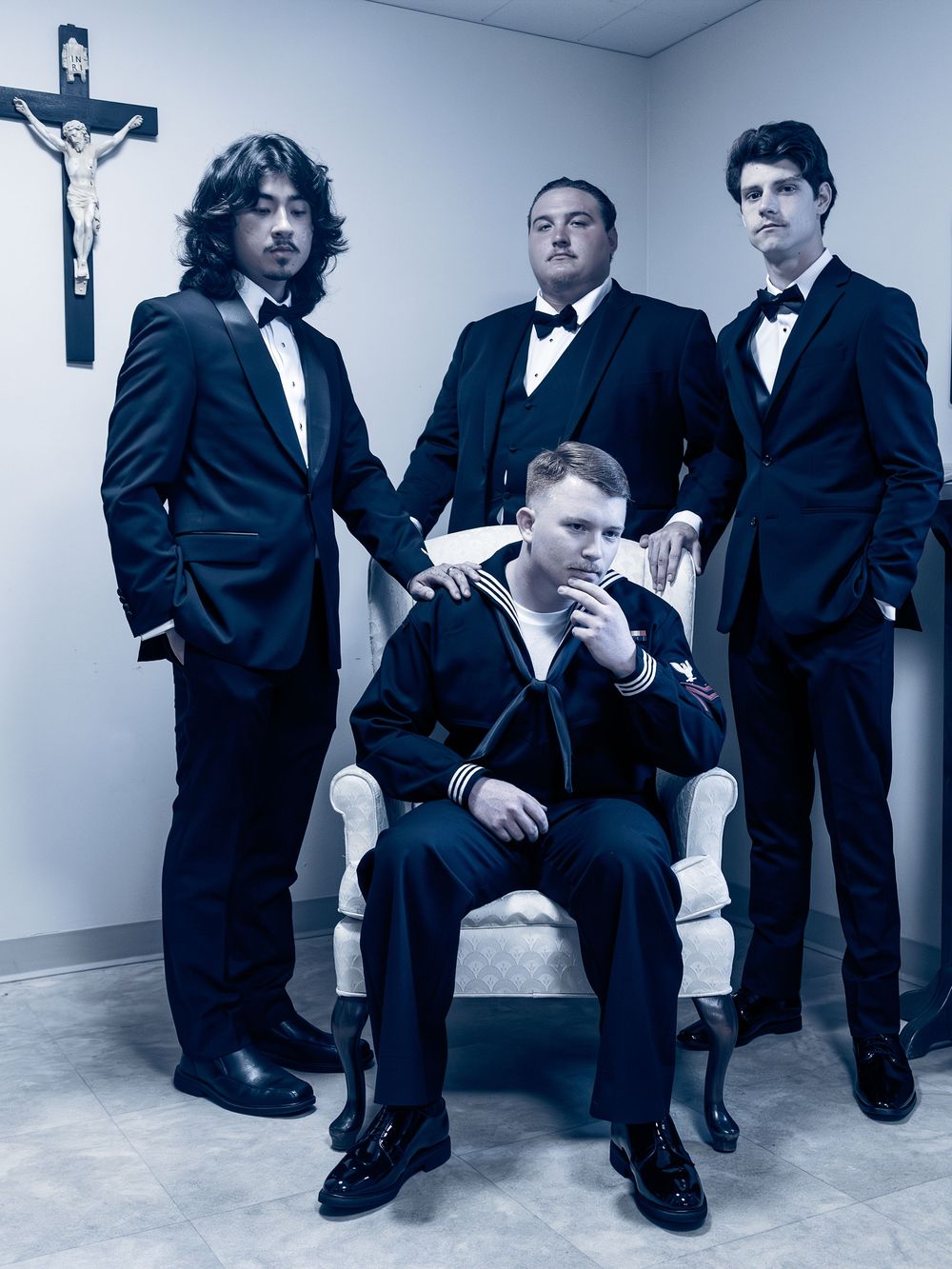 Four men in black tuxedos pose formally in a room with a crucifix, one seated in a white chair wearing a sailor uniform.
