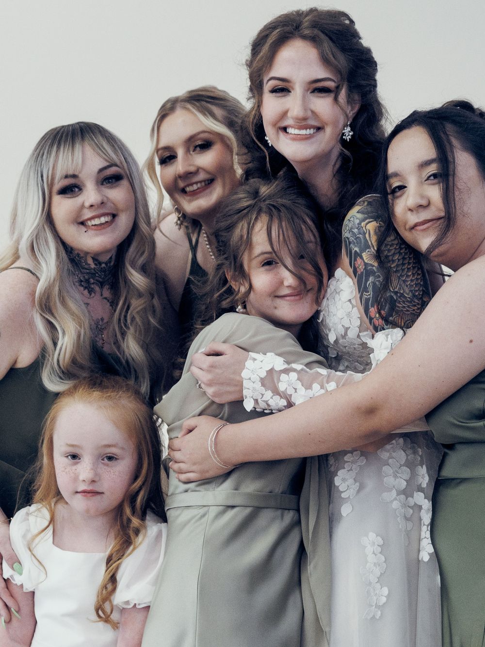 Group of bridesmaids in sage green dresses hugging bride in white floral gown with young flower girl at wedding.