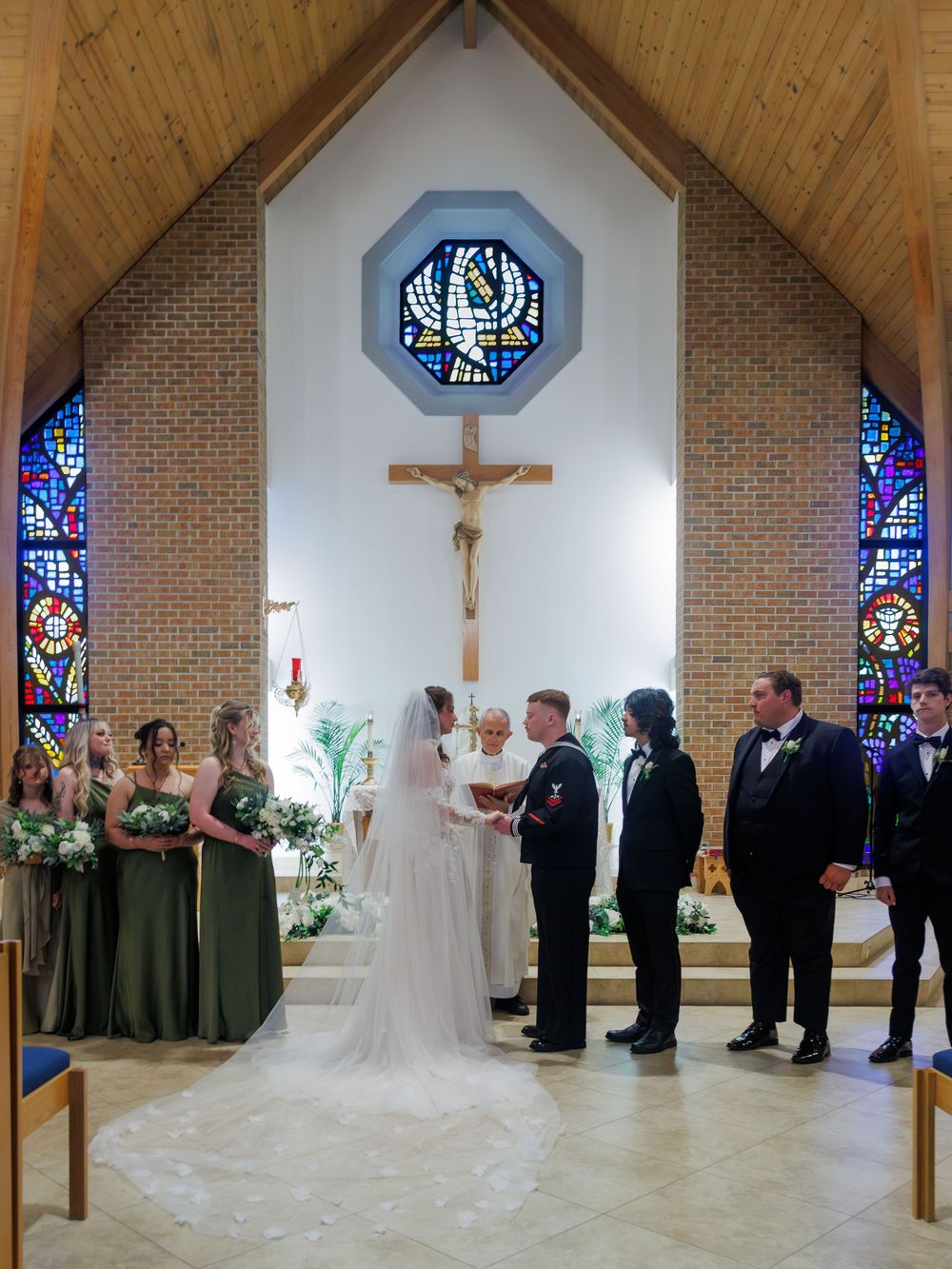 Bride and groom exchange vows at a Catholic church altar surrounded by bridesmaids in green dresses and groomsmen.