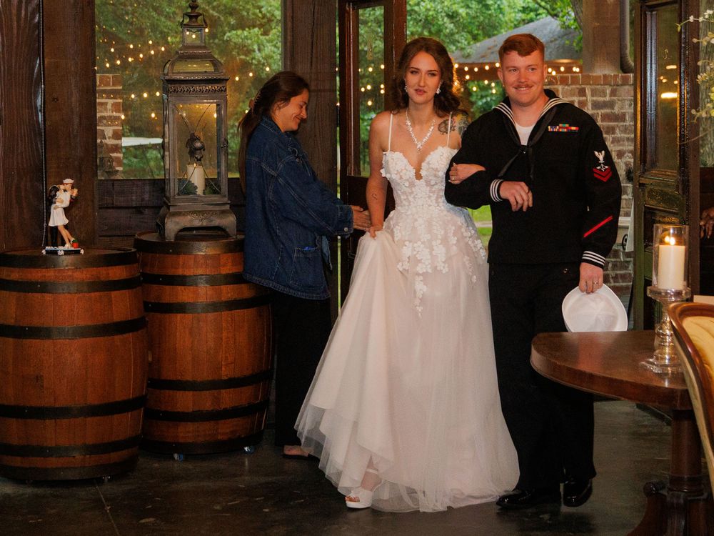 Bride in white gown stands with Navy sailor groom near wooden barrels at rustic wedding venue.