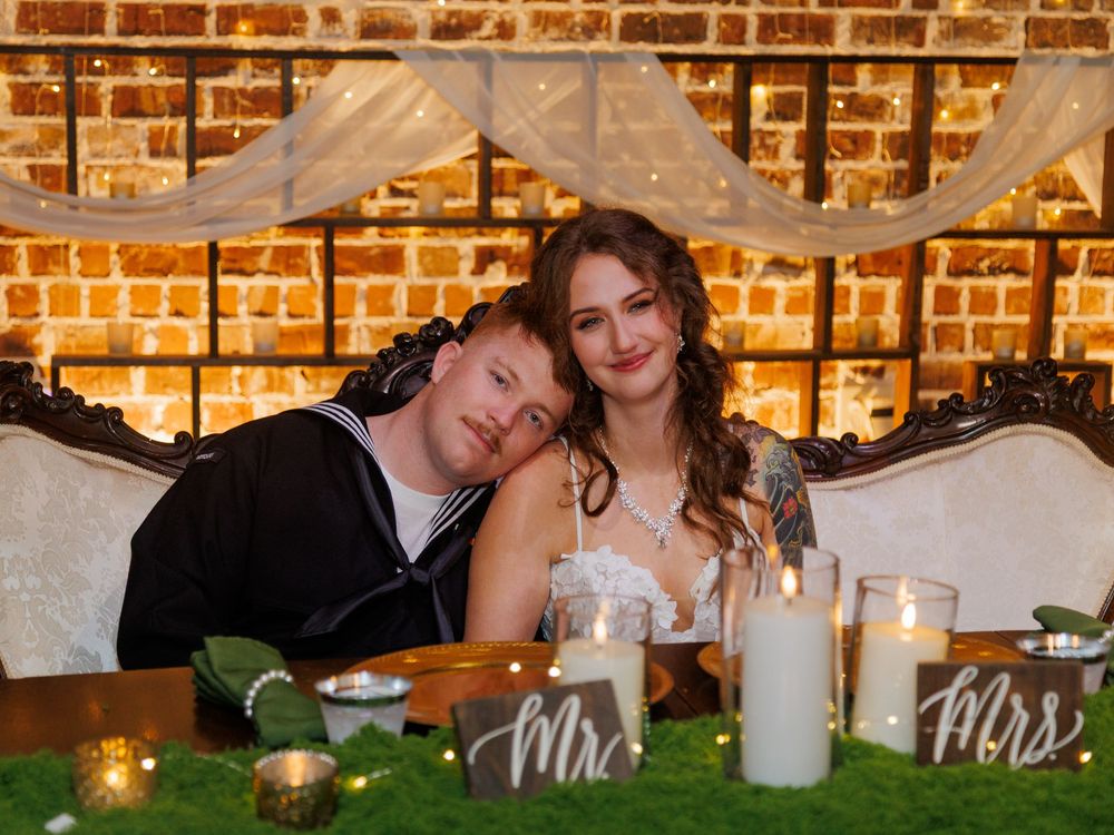 Newlywed couple seated at wedding sweetheart table with Mr. and Mrs. signs, candles, and brick wall backdrop.