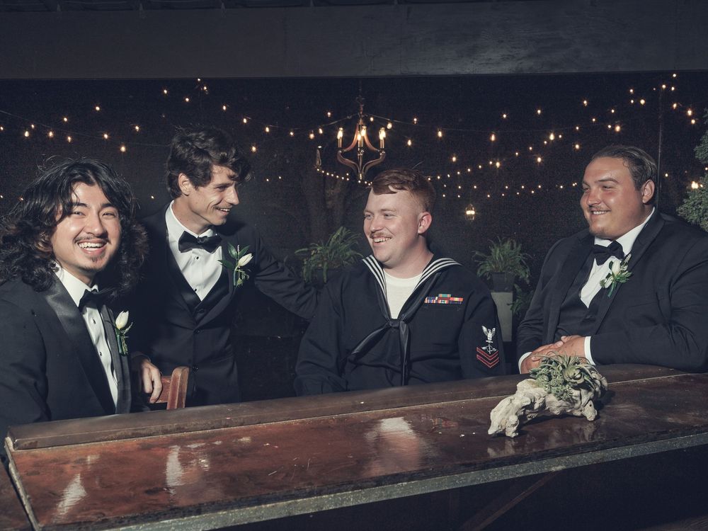 Four young men in formal attire, including one in a Navy uniform, smiling together at a wooden bar with string lights.