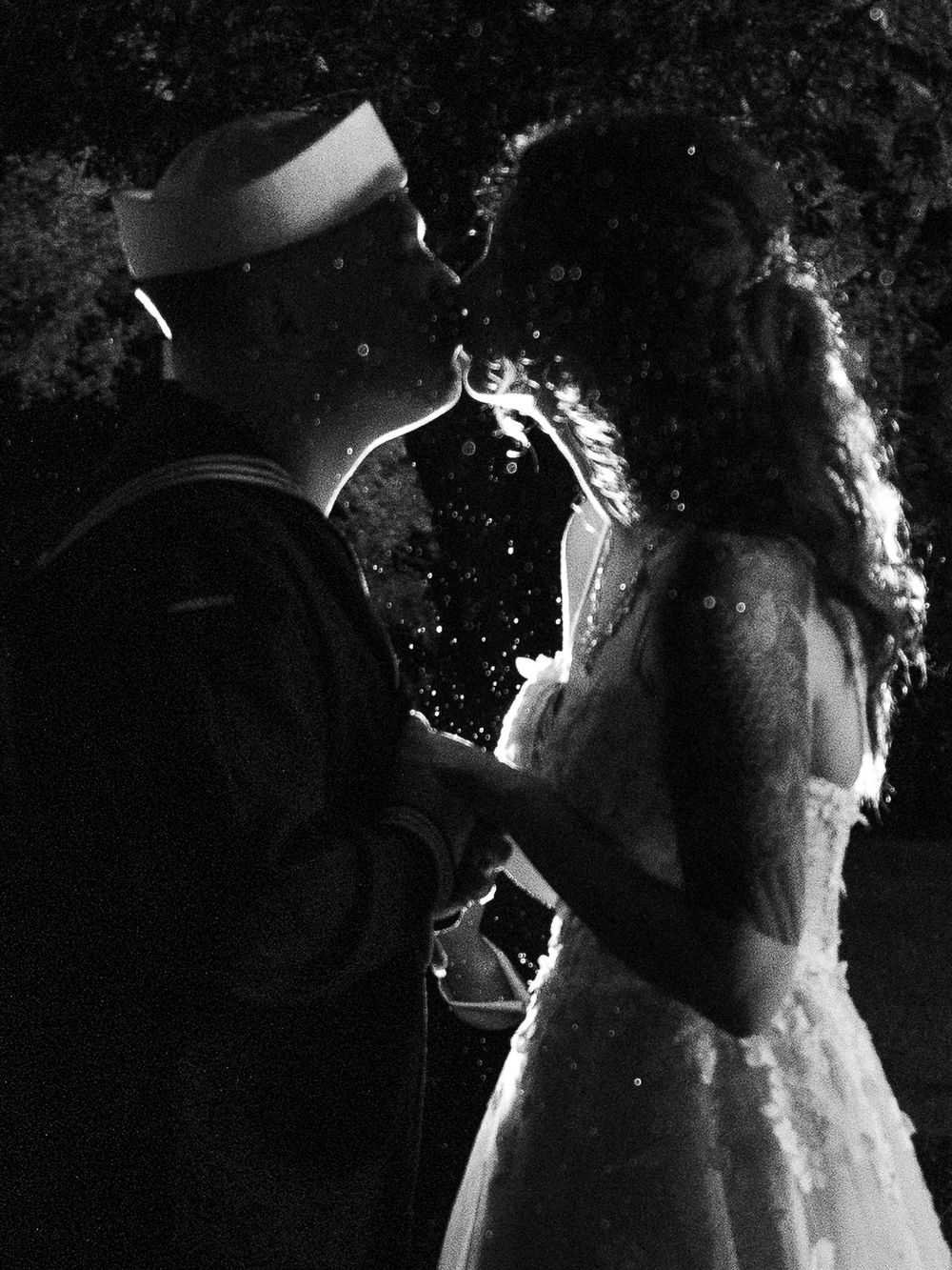 Black and white photo of a Navy sailor and bride sharing a romantic moment in wedding attire at night.