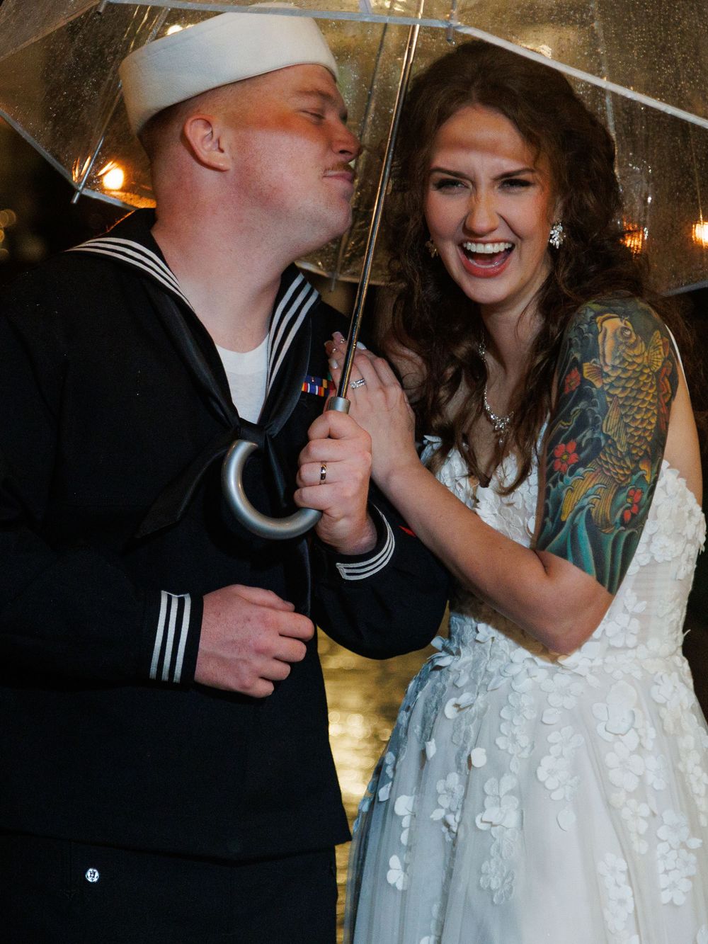Navy sailor and bride share a joyful moment under a clear umbrella on their wedding day, bride wearing a floral gown.