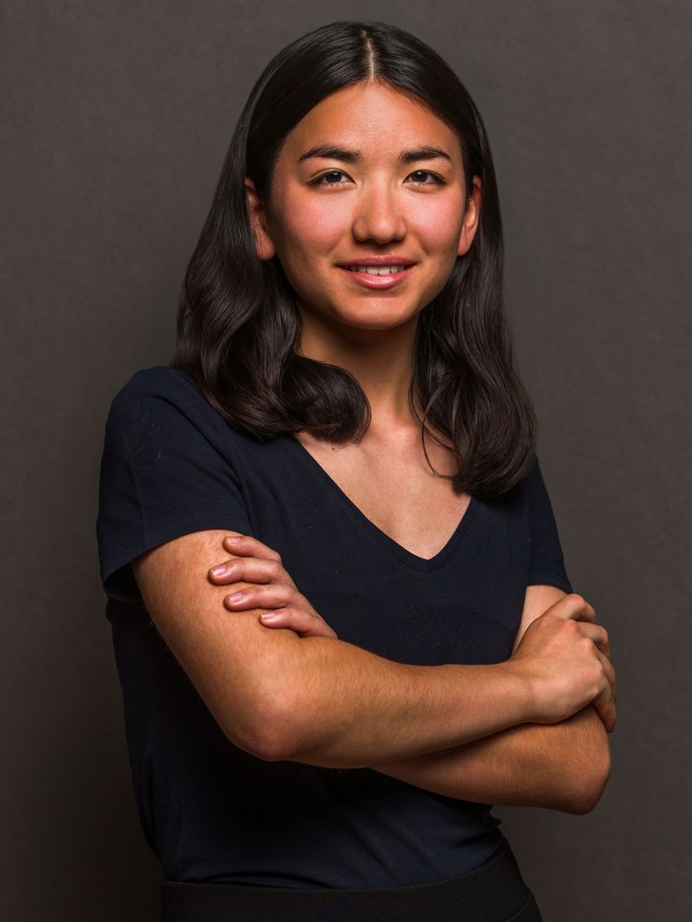 Young woman with dark hair in navy v-neck top, arms crossed, smiling confidently against gray background.