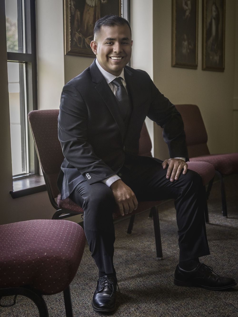 A smiling man in a black suit sits confidently in a chair by a window in a formal indoor setting.