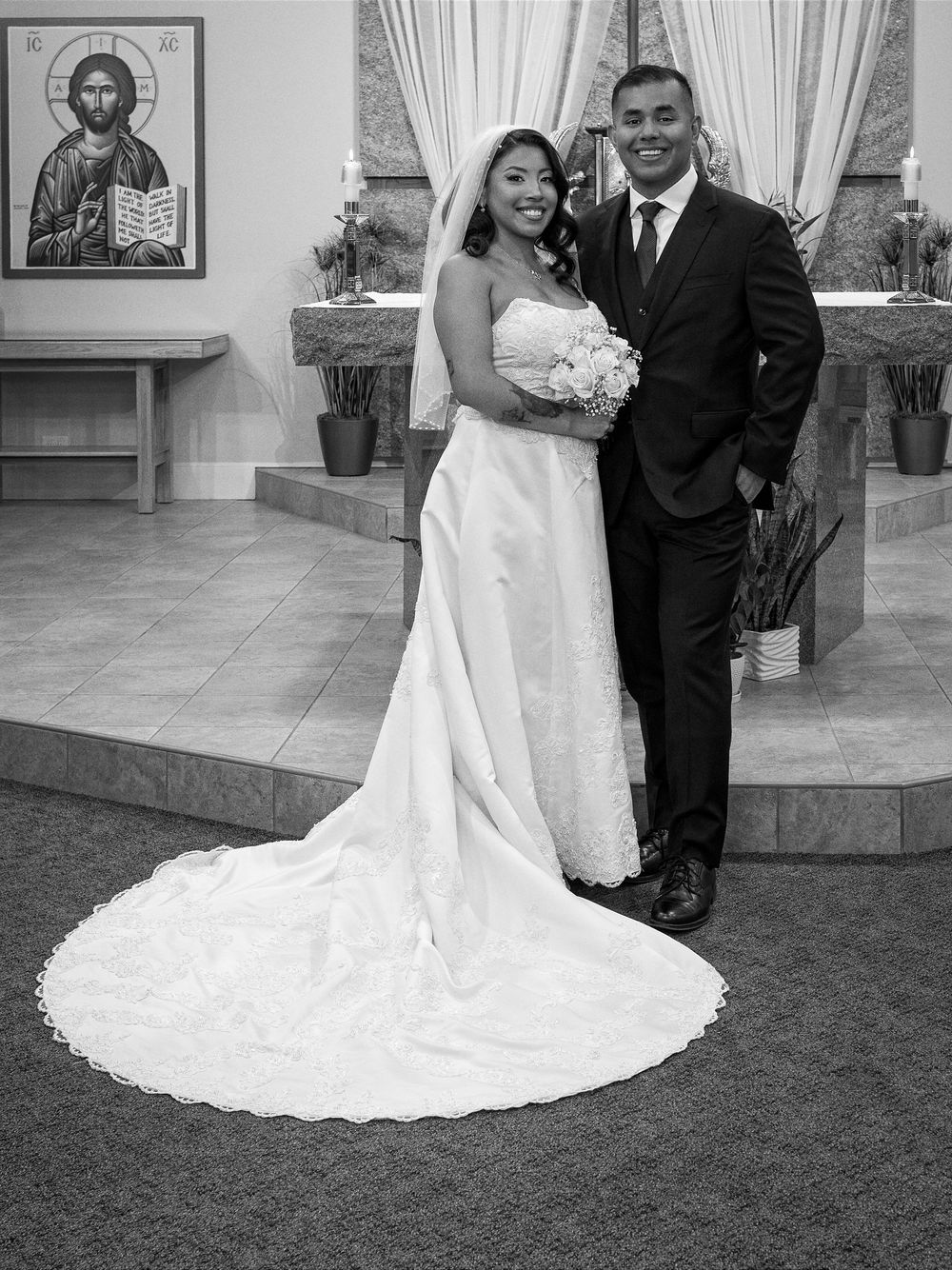 Black and white wedding photo of bride in white gown and groom in suit posing in a church.