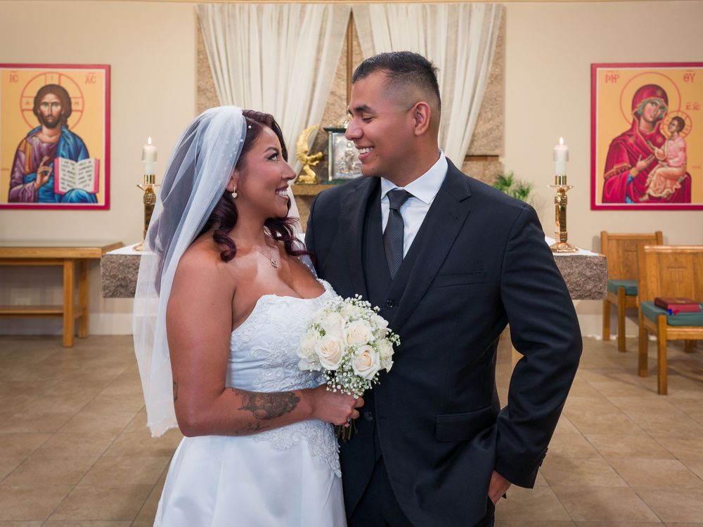 Bride and groom smiling at each other in a church wedding ceremony, bride holding white bouquet, religious icons on wall.