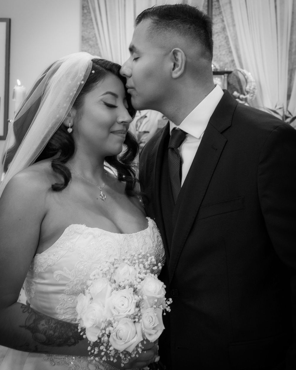Black and white photo of a groom kissing bride on forehead, bride holding white rose bouquet in wedding dress with veil.