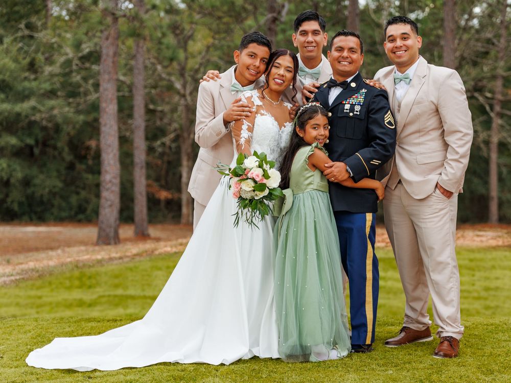 Wedding party poses together outdoors on grassy lawn with pine trees in background during summer celebration.