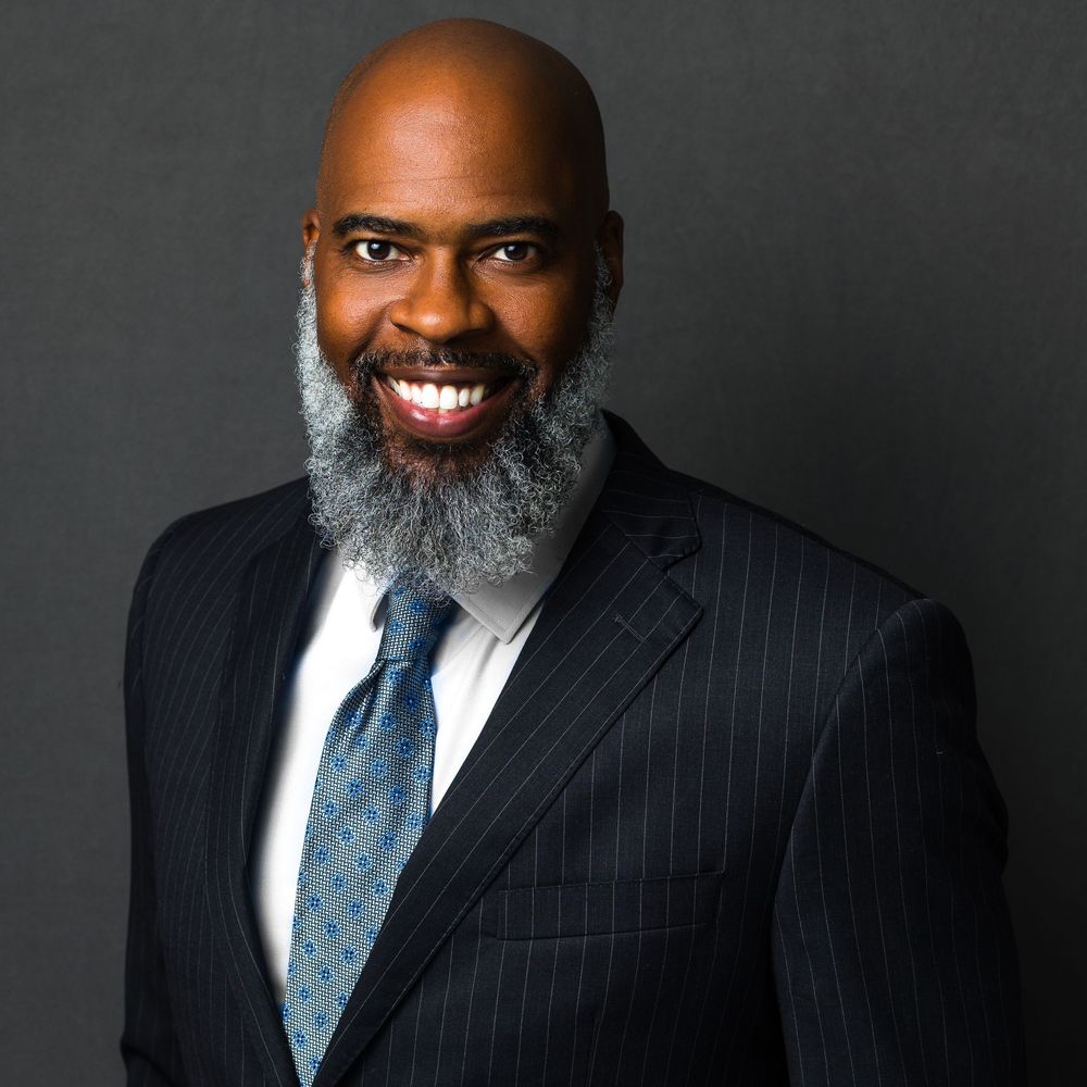 Professional Black man with gray beard wearing navy pinstripe suit and tie, smiling confidently against gray background.