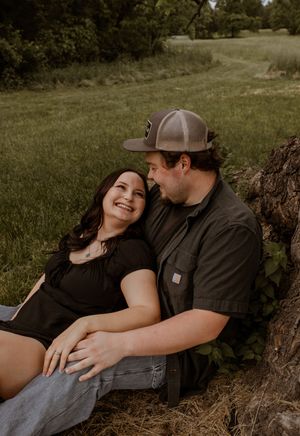 A couple shares an intimate moment together while sitting against a tree trunk in a grassy field.