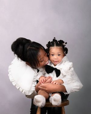 A mother and daughter in white ruffled outfits sitting on a wooden stool against a grey studio backdrop.