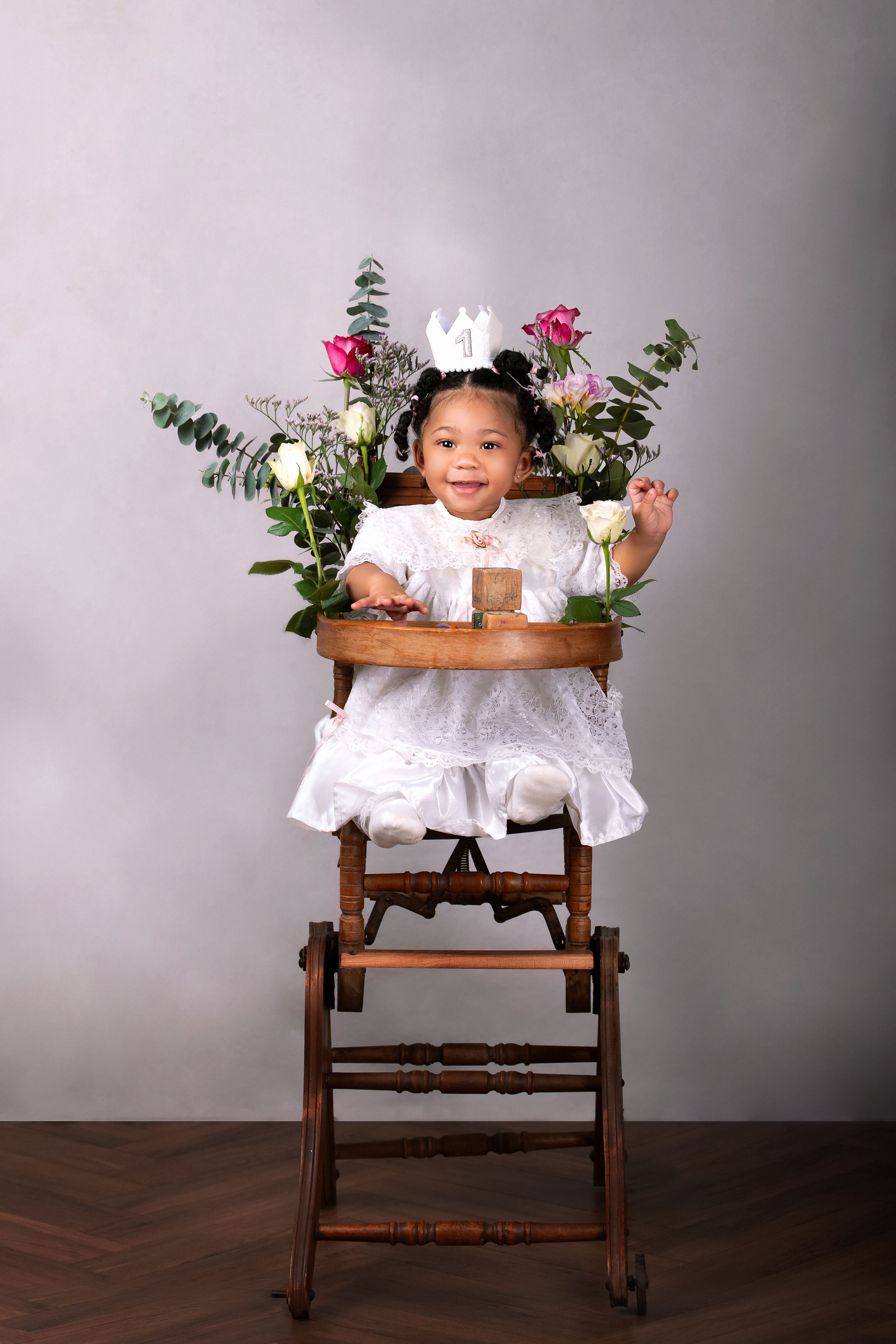 A vintage wooden high chair decorated with white roses and pink flowers on a dark hardwood floor against a grey backdrop with a baby playing
