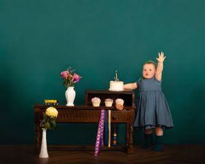 A child in a blue dress celebrates near a small wooden desk with a birthday cake and floral decorations against a teal wall. Heirloom