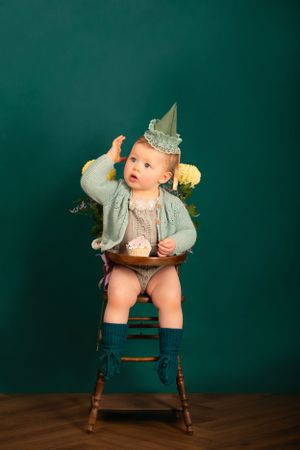 A little one sits on a wooden high chair wearing a mint green cardigan and paper crown against a dark green backdrop.