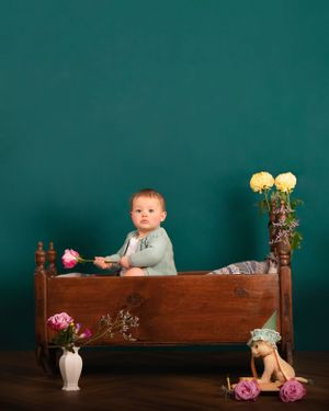 A small child sits in an antique wooden bed with flowers and a teddy bear against a dark teal background. Heirloom nostalgia art