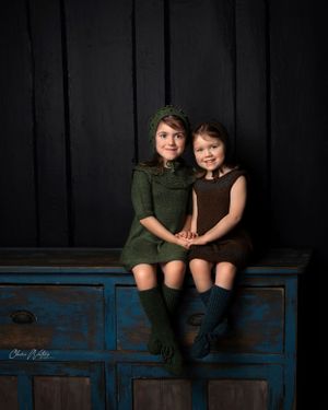 Two children in vintage-style outfits sitting together on a rustic blue cabinet against a black backdrop in a studio setting.