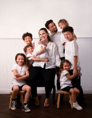 A family group wearing coordinated white shirts poses together on wooden stools against a light-colored wall.
