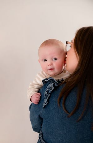 Baby peering over Mum's shoulder as his Mum gives him kisses on the check
