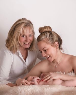 Mums & Grandmother share a joyful moment together while sitting on soft white fur, with Newborn baby creating an intimate portrait setting.