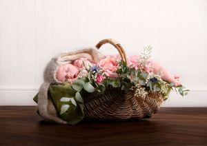 Newborn baby sleeping peacefully in wicker basket decorated with pink flowers and green foliage.