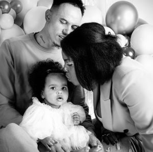 Black and white portrait of Jamaican Mama kissing her baby girl, while father hugs them both surrounded by celebrations