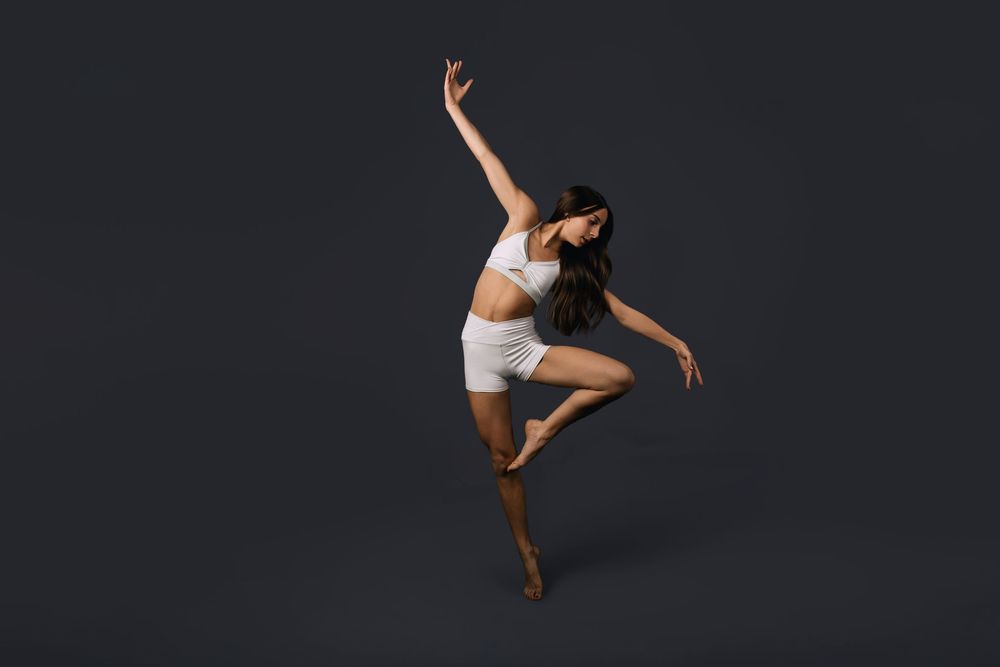 A female dancer leaps gracefully in white shorts and crop top against a dark gray studio background.