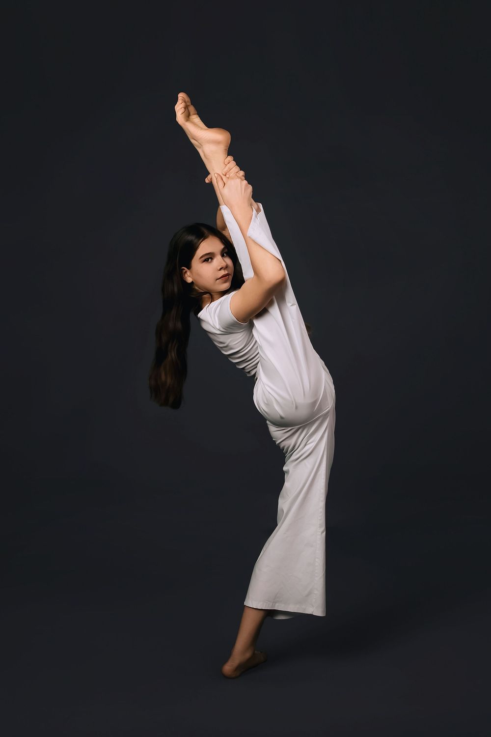 A flexible young woman in white outfit performs a standing split pose against a dark background.